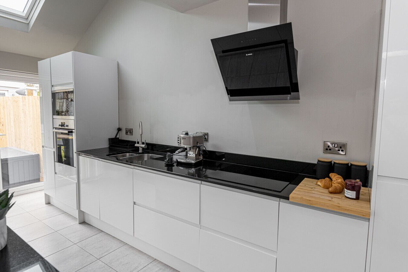 Una cocina moderna con armarios blancos, encimera negra, horno empotrado, cafetera y tabla de madera con un croissant y mermelada.