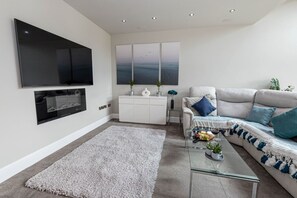 A modern living room with a flat-screen TV, a white sofa with blue cushions, a glass coffee table, and a large painting above the sofa. - Sarnia Retreat - Open plan living area & garden close to Poole Quay (Poole)