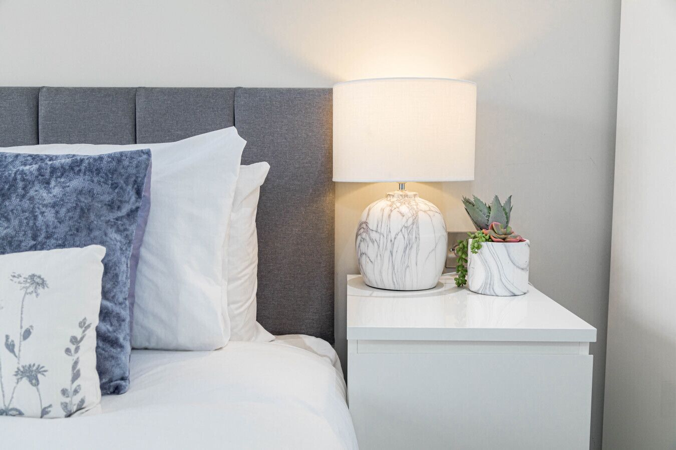 En pænt redt seng med hvide og blå puder, et gråt sengegavl, et natbord med en lampe og en potteplante.