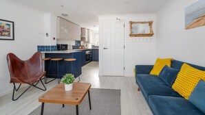 A modern living room with a blue sofa, a brown leather chair, a wooden coffee table, and a kitchen in the background. - Driftwood - Garden & parking close to beaches and town (Dorset)