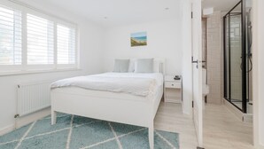 A bedroom with a bed, a nightstand, a painting, a bathroom with a glass shower, and a view of the outside. - Driftwood - Garden & parking close to beaches and town (Dorset)