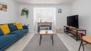 A modern living room with a blue sofa, a coffee table, a TV, and a window with blinds. - Driftwood - Garden & parking close to beaches and town (Dorset)