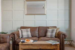 A brown leather sofa with plaid cushions, a wooden coffee table with a cheese board and magazine, and a wall-mounted mirror. - Ocean Spray Suite - Within moments of beaches, the seafront & town (Bournemouth)
