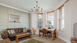 A living room with a brown sofa, a wooden coffee table, a dining table with chairs, a chandelier, and large windows with blinds. - Ocean Spray Suite - By the beaches, the seafront & town (Bournemouth)