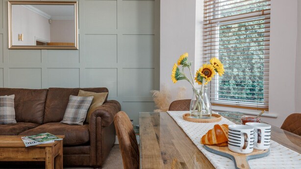 A living room with a brown leather sofa, a wooden coffee table, a dining table with a vase of sunflowers, and a window with blinds. - Ocean Spray Suite - By the beaches, the seafront & town (Bournemouth)