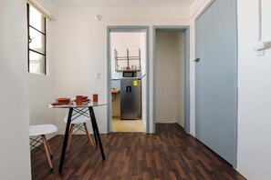 Un couloir étroit mène à un espace repas agrémenté d’une table et de chaises en verre, puis à une cuisine équipée d’un réfrigérateur et d’un four à micro-ondes, et enfin à une porte qui donne sur une autre pièce.