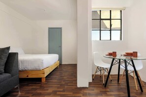 Une chambre à coucher avec un lit, un plancher en bois, un canapé gris, une table en verre avec des chaises et une fenêtre avec des stores.