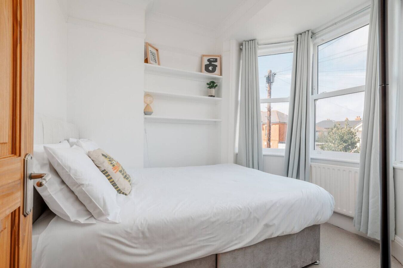 Une chambre à coucher avec un lit, une fenêtre avec des rideaux, une étagère avec des objets décoratifs et une vue sur l’extérieur.