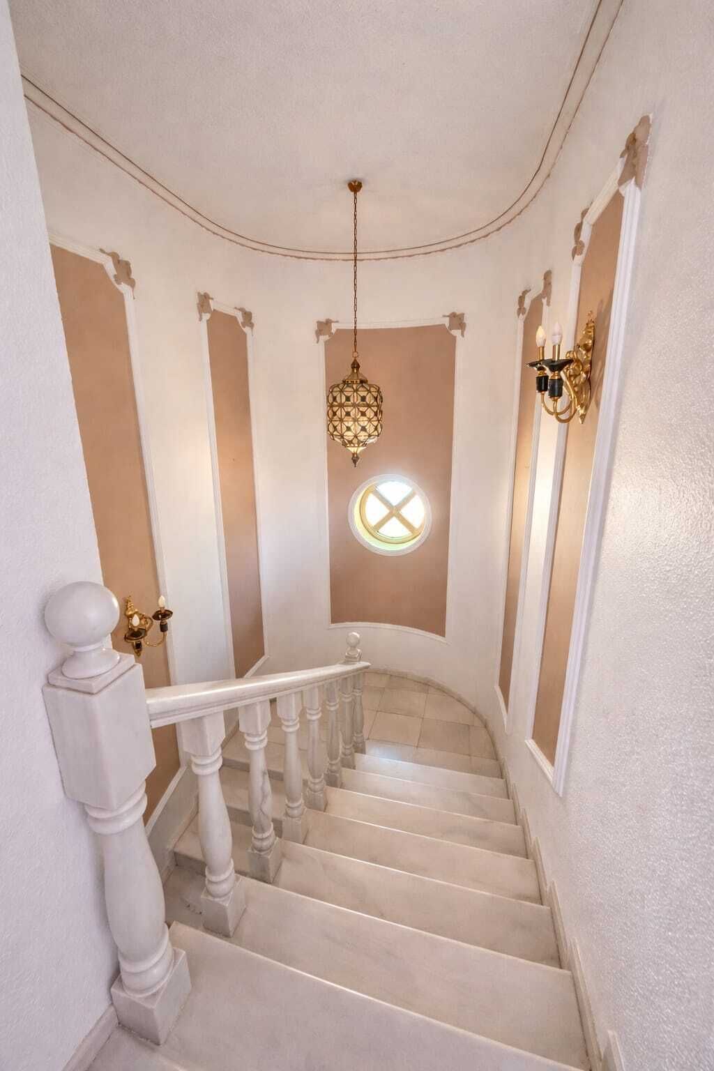 Eine Treppe mit weißen Balustraden, einem runden Fenster mit Kreuzmuster und einem hängenden Laternenleuchter.