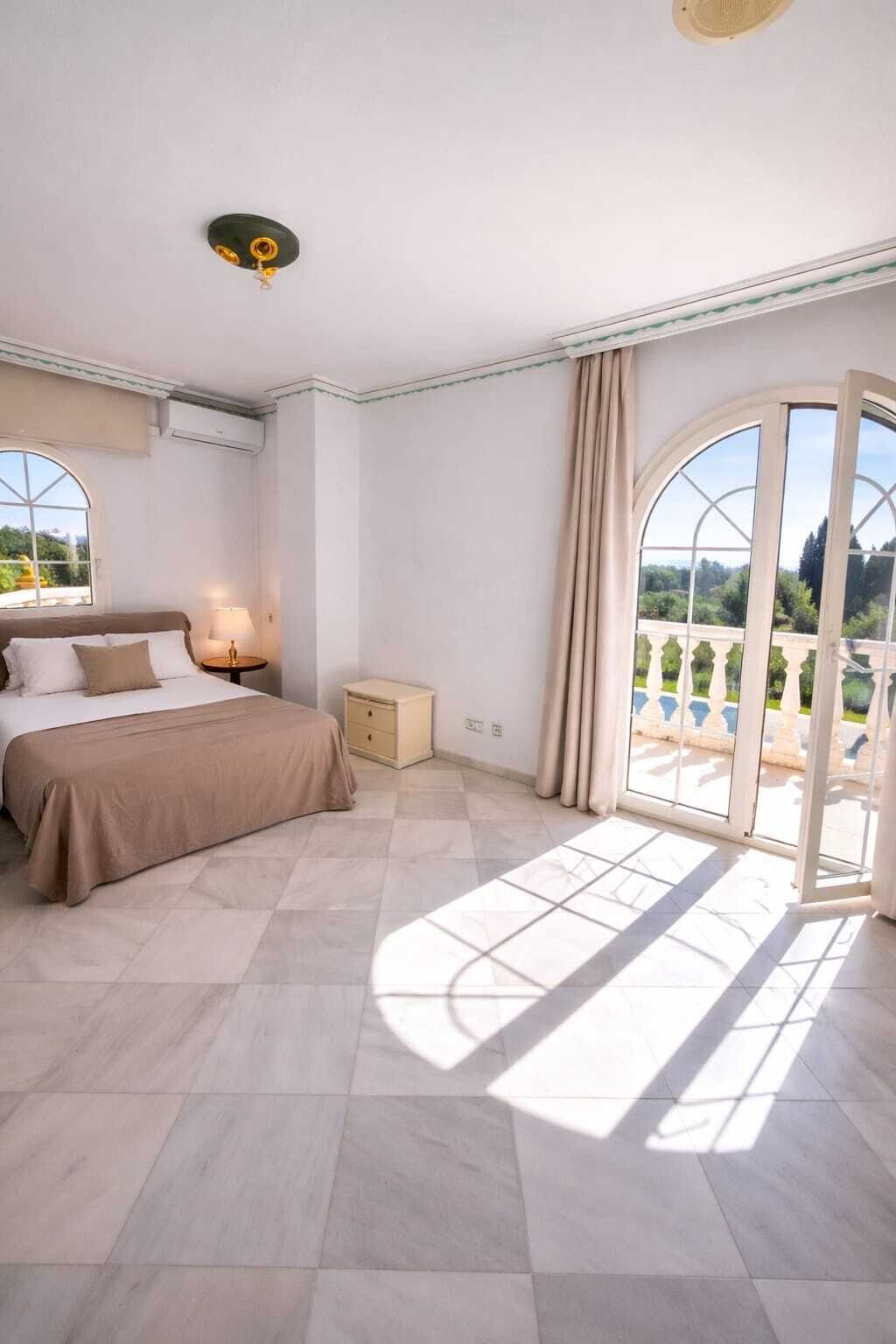 A bedroom with a bed, bedside table, and a view of a garden through a large window.