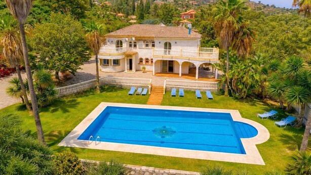 Ett stort hus med en swimmingpool, omgivet av palmer och ett kuperat landskap.