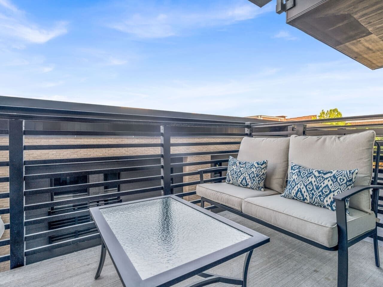 A rooftop patio with a glass table and a sofa with patterned cushions.