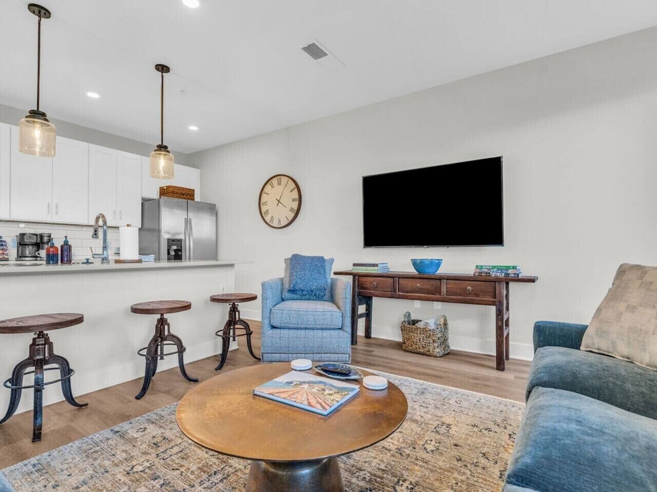 A modern living room with a coffee table, a blue armchair, a sofa, a TV, and a kitchen area with bar stools.
