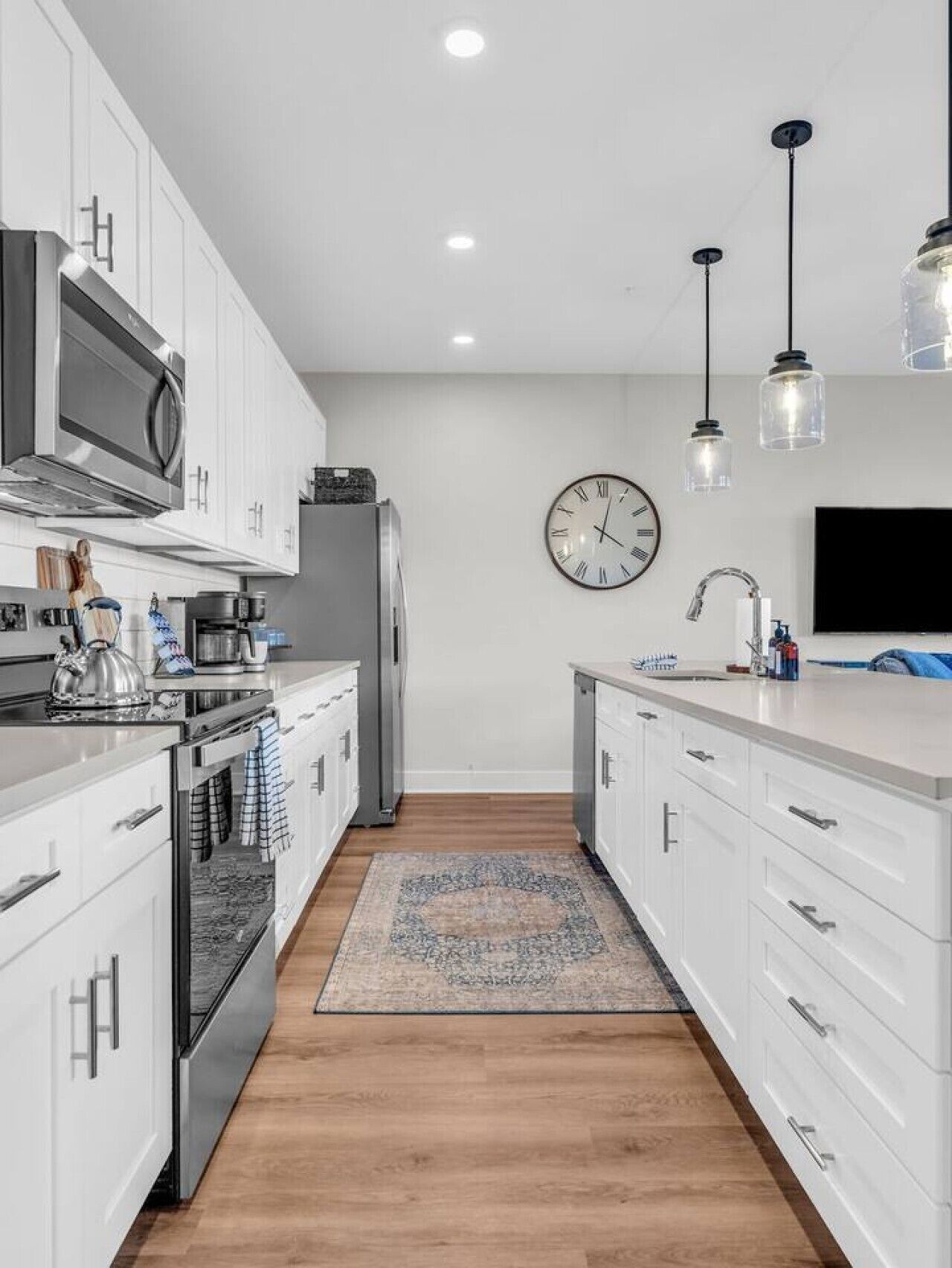 Une cuisine moderne avec des meubles blancs, des appareils en acier inoxydable, une horloge, des luminaires suspendus et un plancher en bois.
