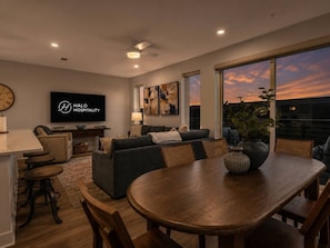 A modern living room with a dining area, a flat-screen TV, and a large window with a sunset view. - Lone Tree Pass | King + Queen | Balcony + Parking (Knoxville)