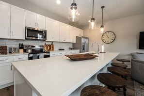 A modern kitchen with white cabinets, stainless steel appliances, a large island with stools, and pendant lighting. - Lone Tree Pass | King + Queen | Balcony + Parking (Knoxville)