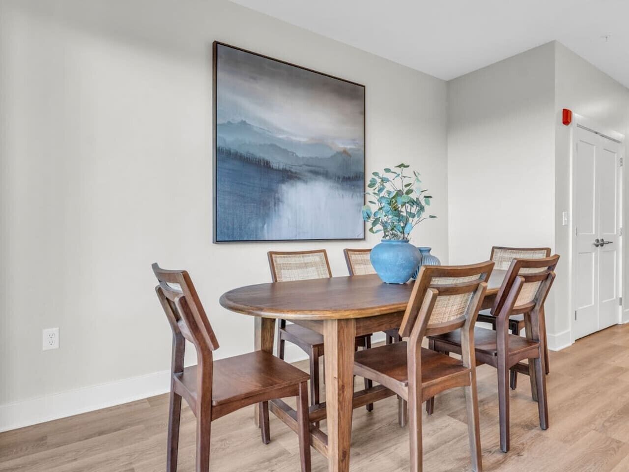 A dining area with a round wooden table and six chairs, a large abstract painting on the wall, and a potted plant.