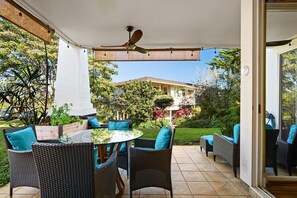 Eine Terrrasse im Freien mit Korbmöbeln, einem Deckenventilator und Blick auf ein Haus und einen Garten.
