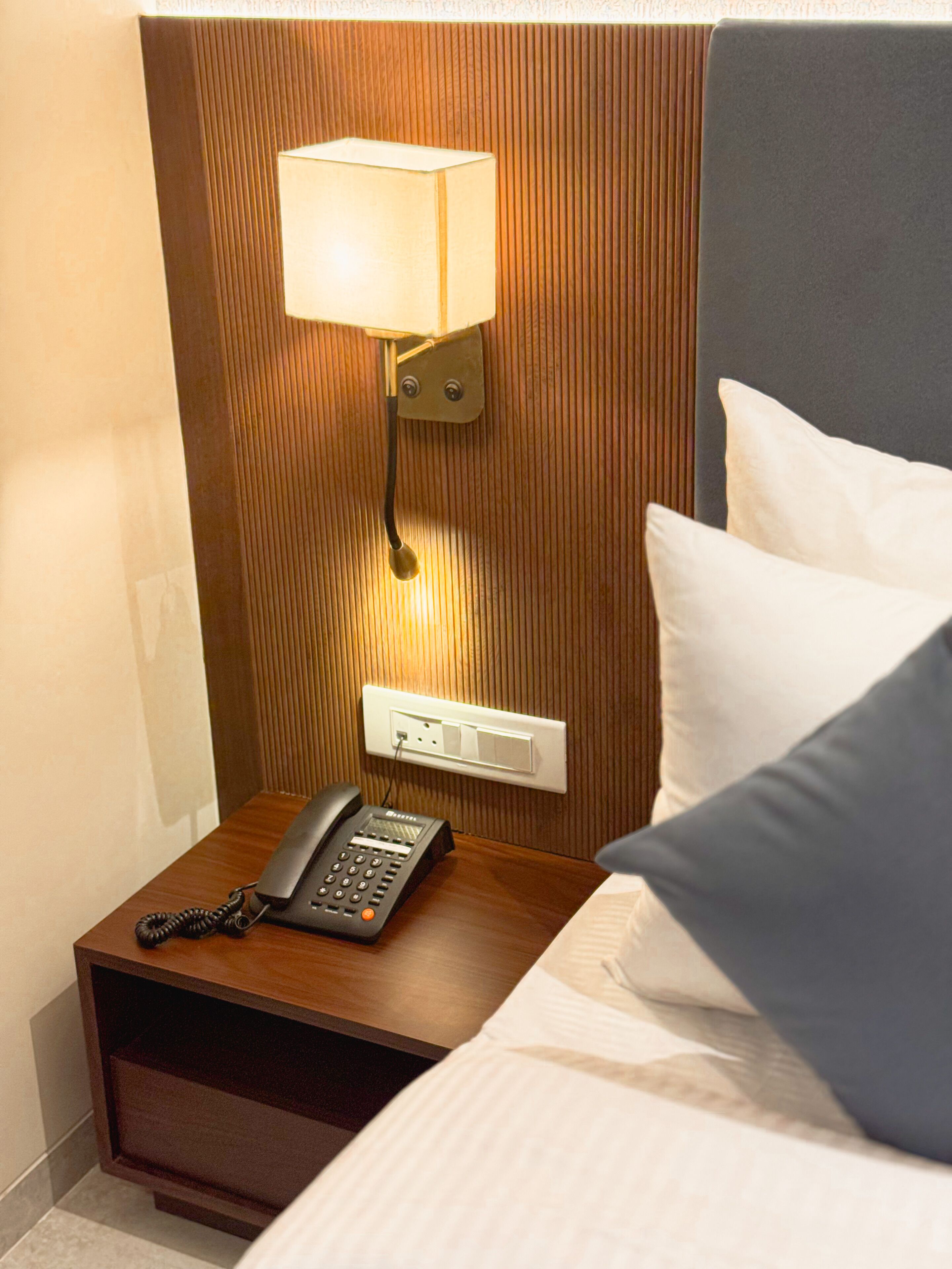 A hotel room with a wooden headboard, a bedside lamp, a telephone, and a bed with pillows.