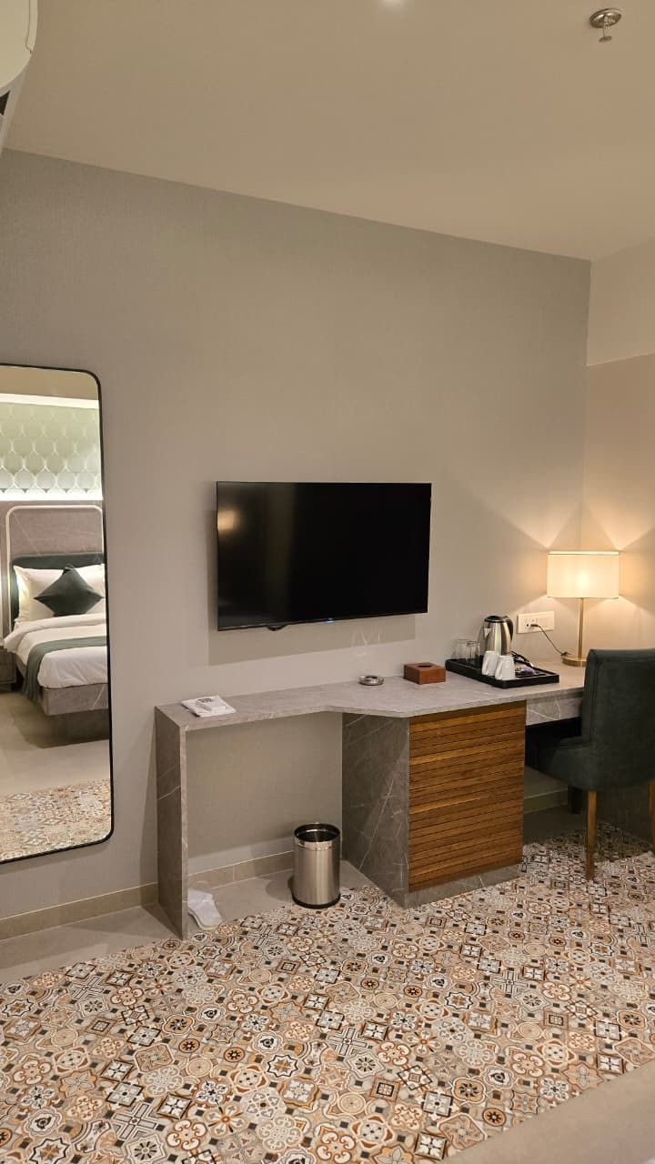 A modern hotel room with a flat-screen TV, a desk with a lamp, a chair, and a patterned floor.