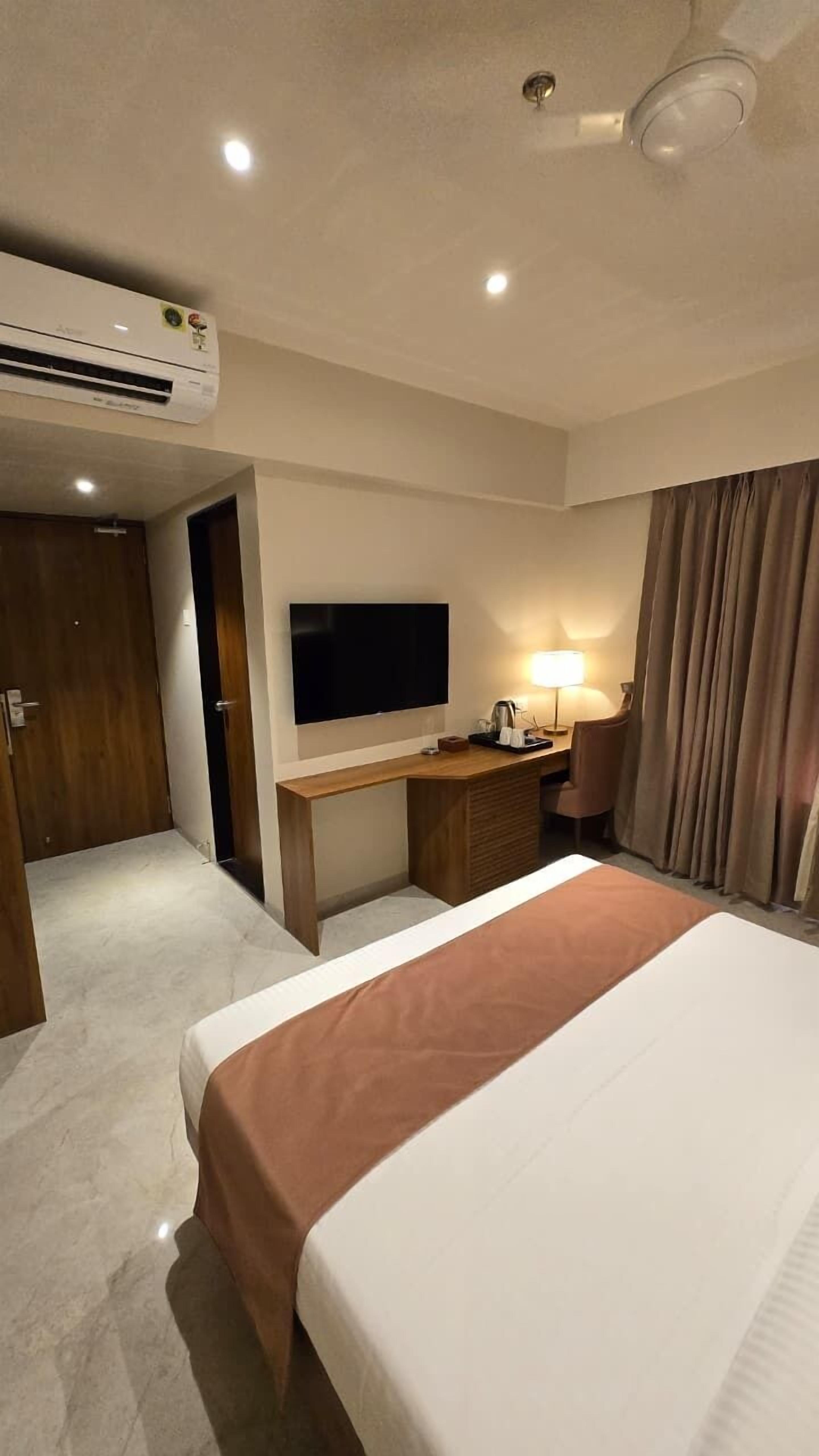A hotel room with a bed, a desk, a television, an air conditioning unit, and a door.