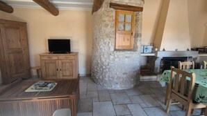 A rustic interior with a stone pillar, wooden furniture, a television on a cabinet, and a stone fireplace. - Le Puits des Oliviers (Puimoisson)