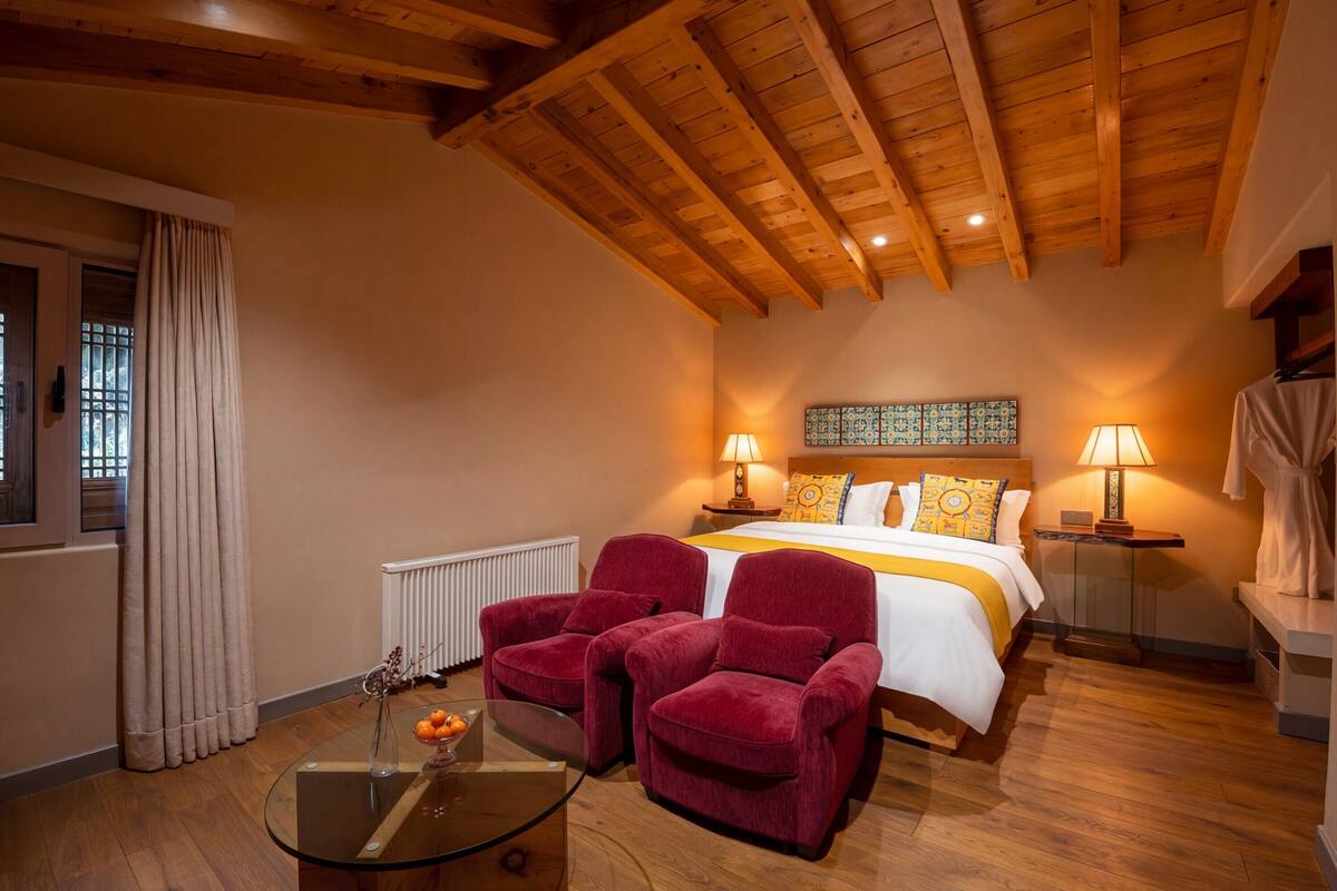 Una habitación de hotel con techo de madera, una cama con ropa de cama blanca, dos sillones rojos, una mesa de centro de vidrio y un radiador.
