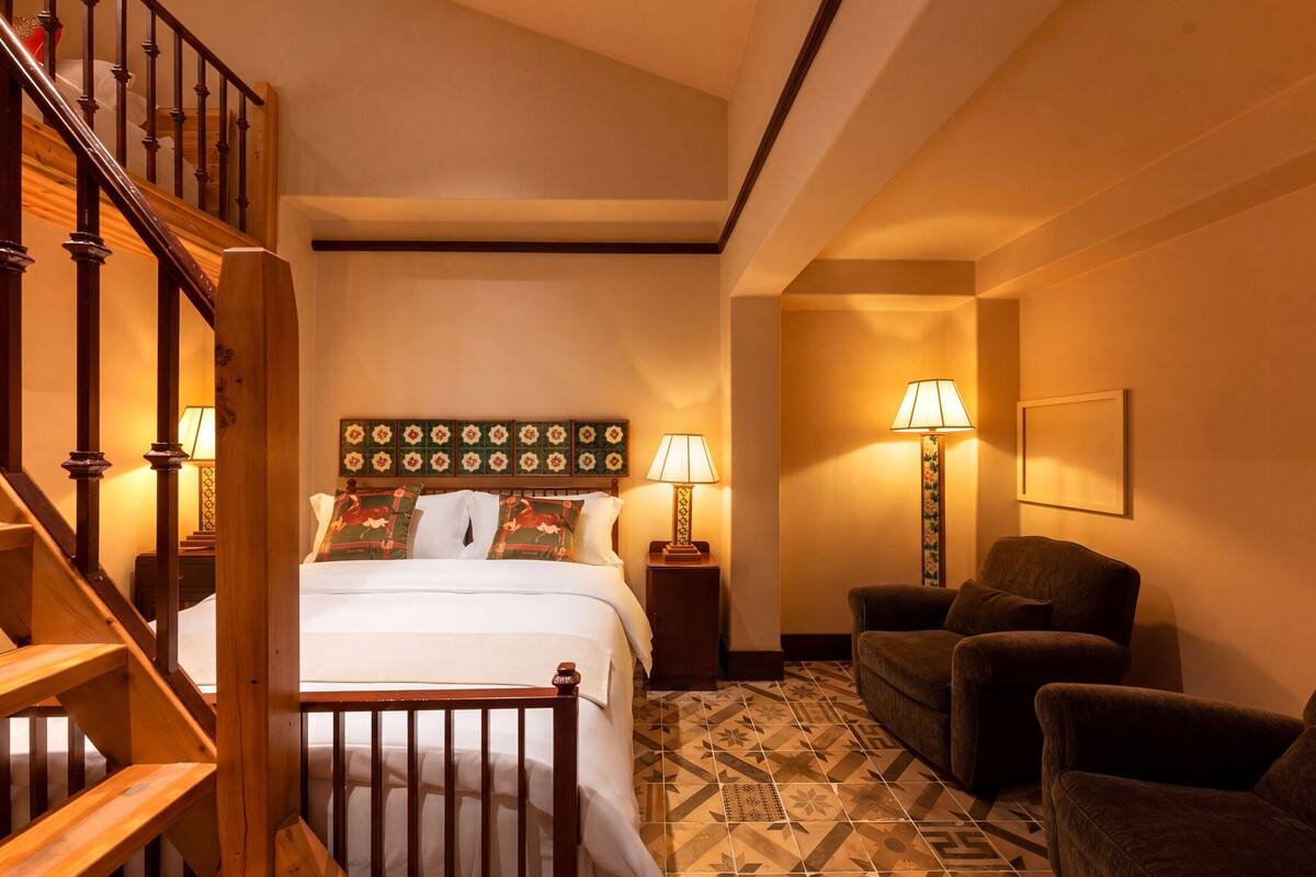 Habitación de hotel con escalera de madera, cama con cabecera floral, dos lámparas de noche, sofá y baldosas de suelo estampadas.