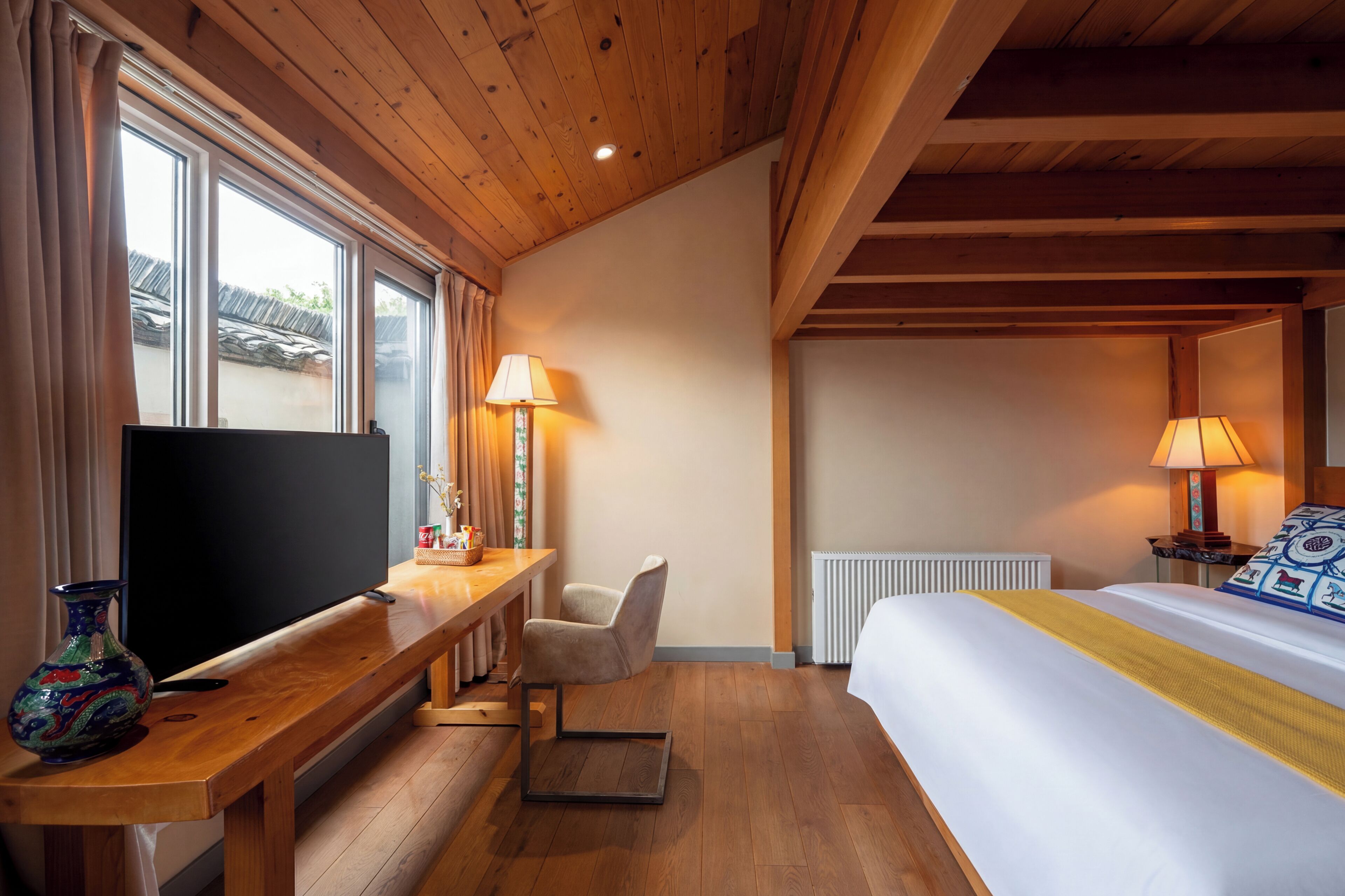 A hotel room with a wooden ceiling, a bed with a yellow bedspread, a TV on a wooden table, a chair, and two lamps.