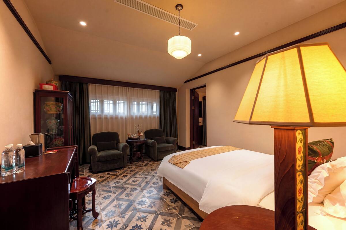 Habitación de hotel con una cama, un escritorio con silla, una mesa con sillas, una lámpara y una ventana con cortinas.