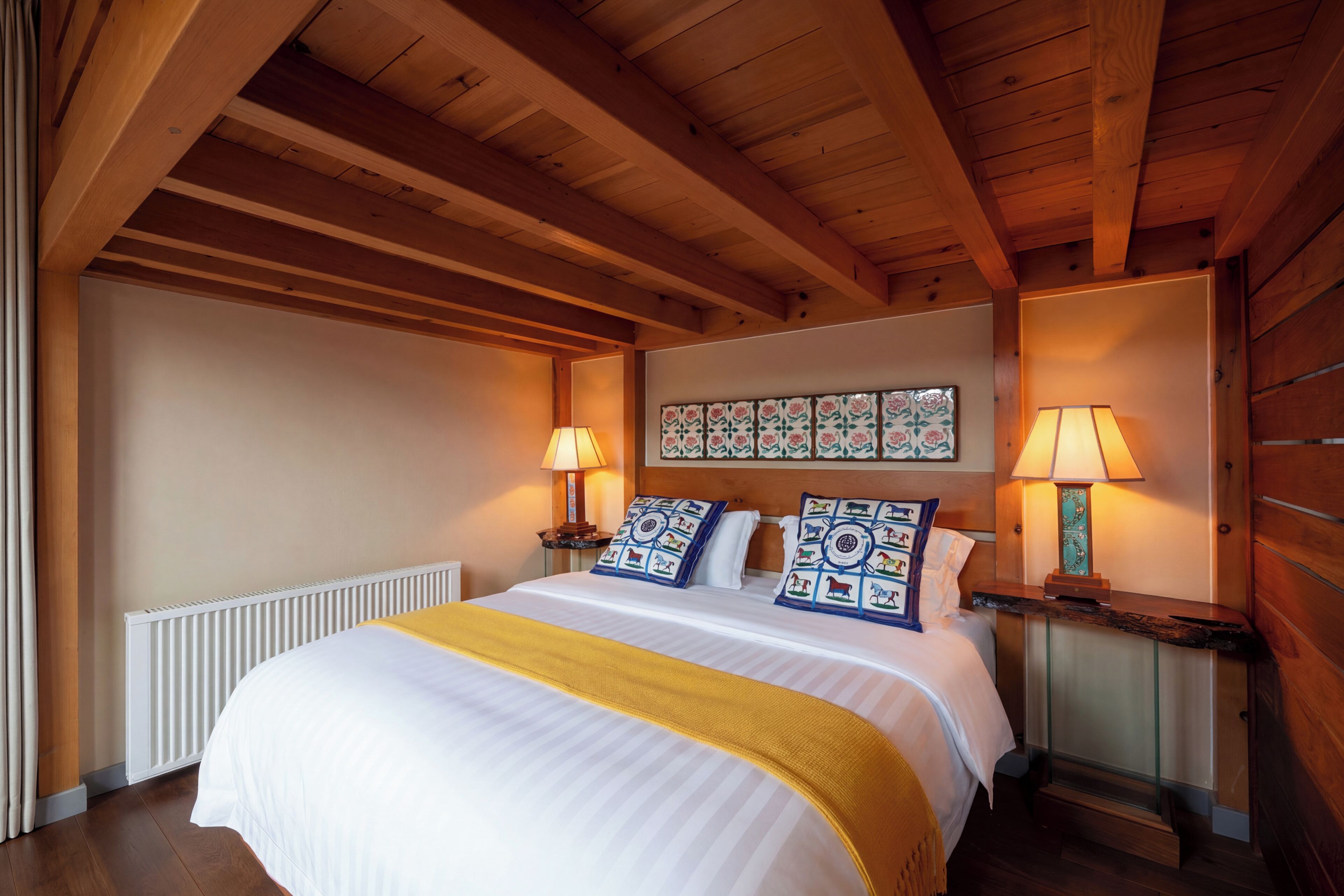A bedroom with a wooden ceiling, a bed with white bedding and a yellow throw, two bedside tables with lamps, and a wall with a floral-patterned artwork.