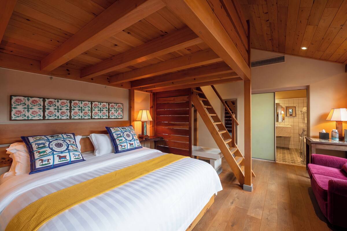Un dormitorio con cama, vigas de madera, una escalera y un baño visible a través de una puerta abierta.