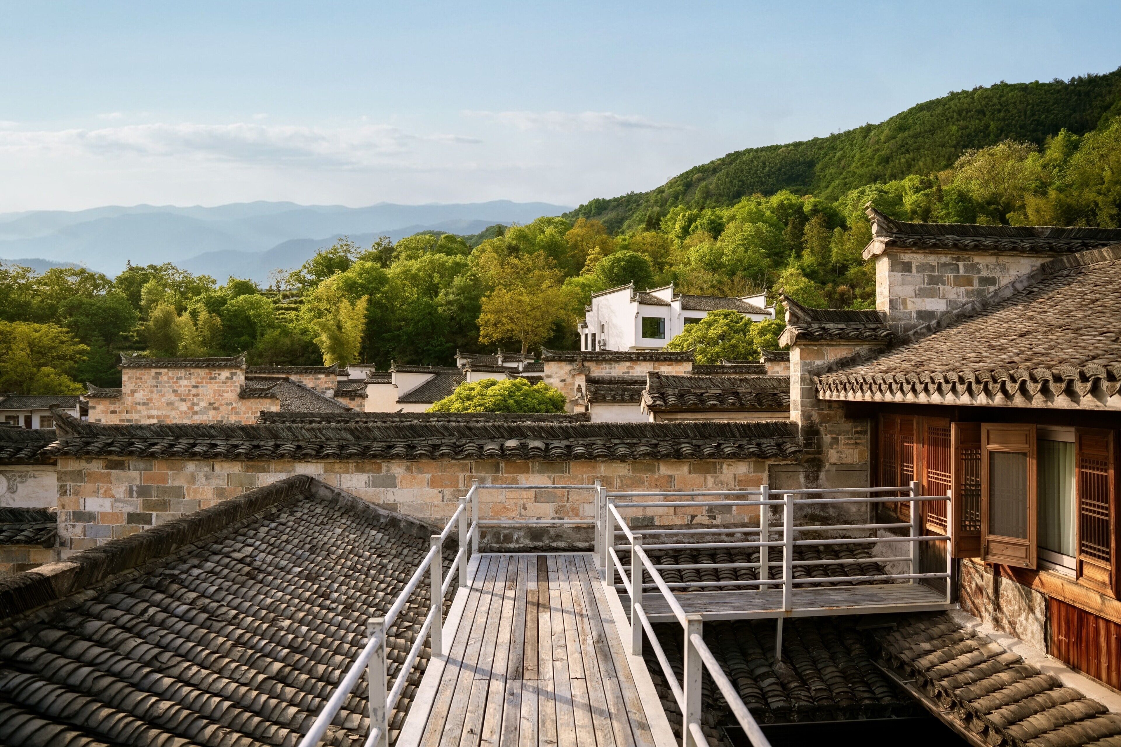 A traditional building with a wooden balcony, stone walls, and a view of a forested hillside.