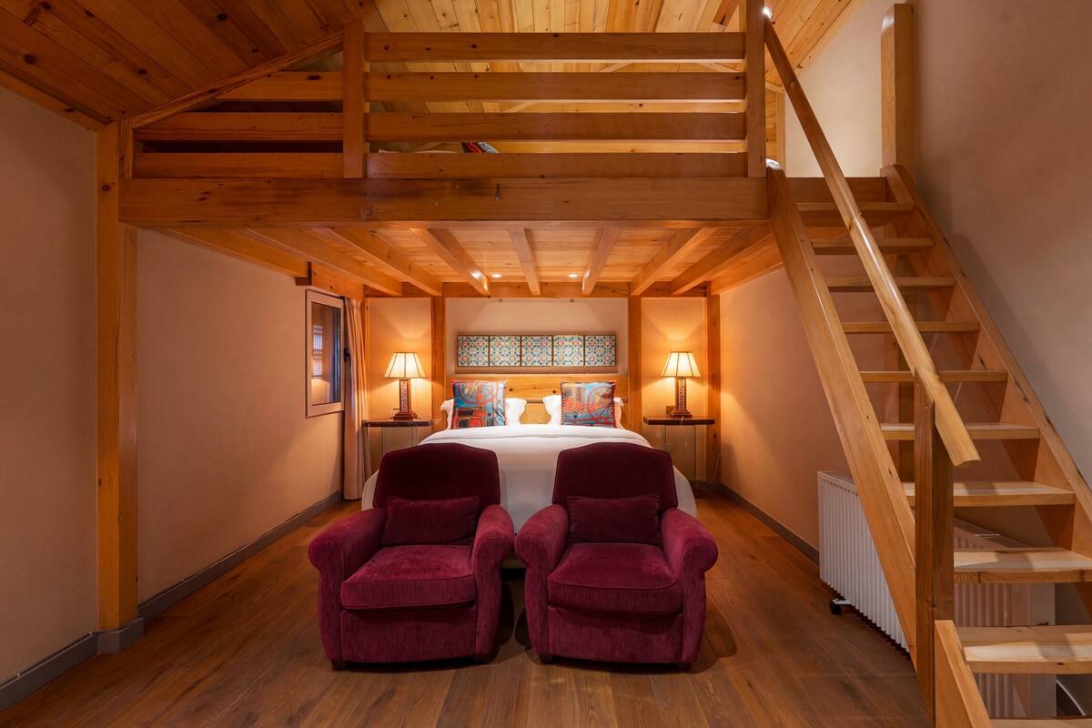 Un dormitorio con techo de madera, una cama con edredón blanco, dos sillones rojos, una mesita de noche con lámpara y una escalera de madera.