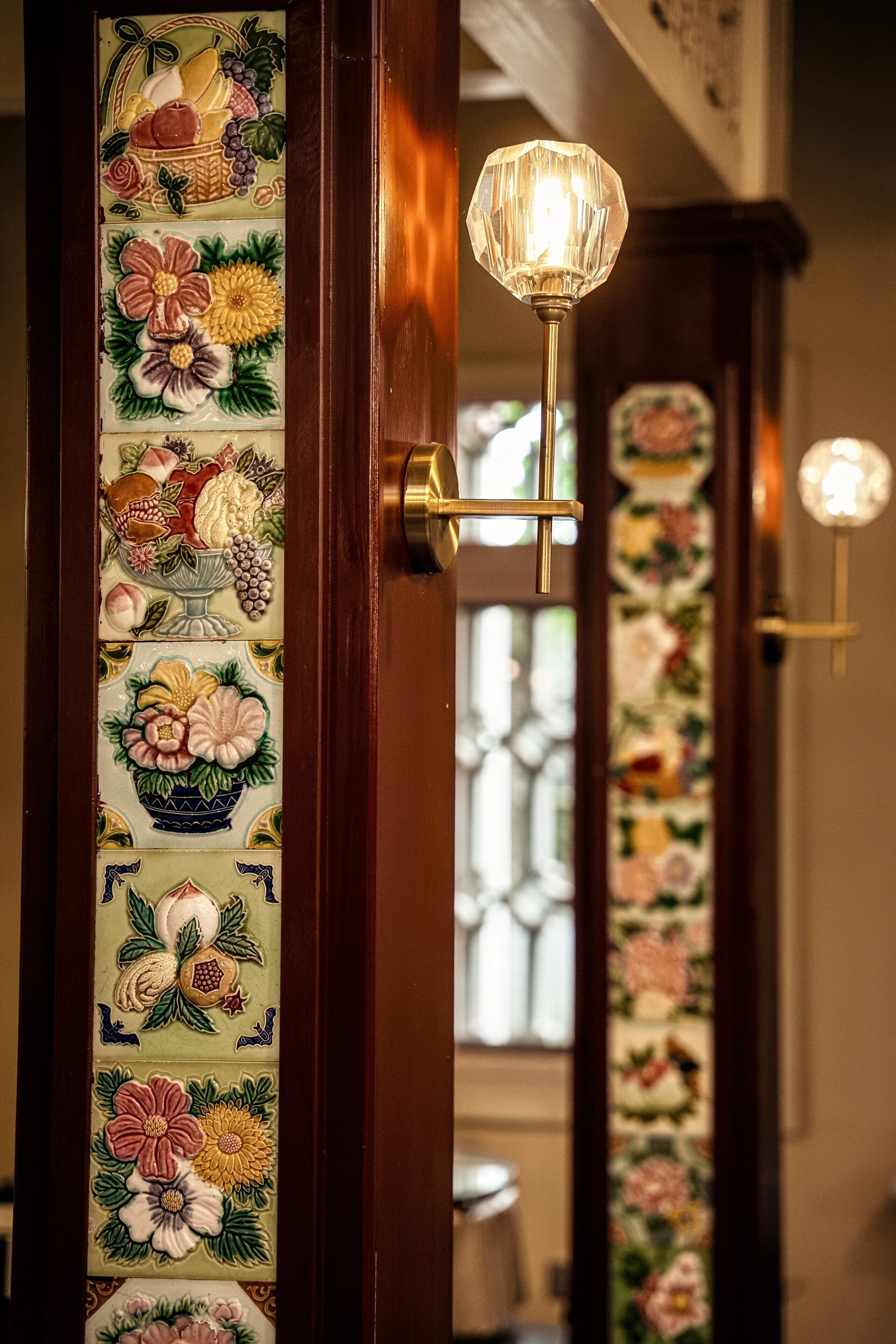 Puerta con un panel decorativo de azulejos con motivos florales y frutales, un picaporte de bronce y una lámpara fijada a la pared.