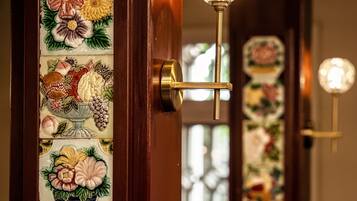 Puerta con un panel decorativo de azulejos con motivos florales y frutales, un picaporte de bronce y una lámpara fijada a la pared.