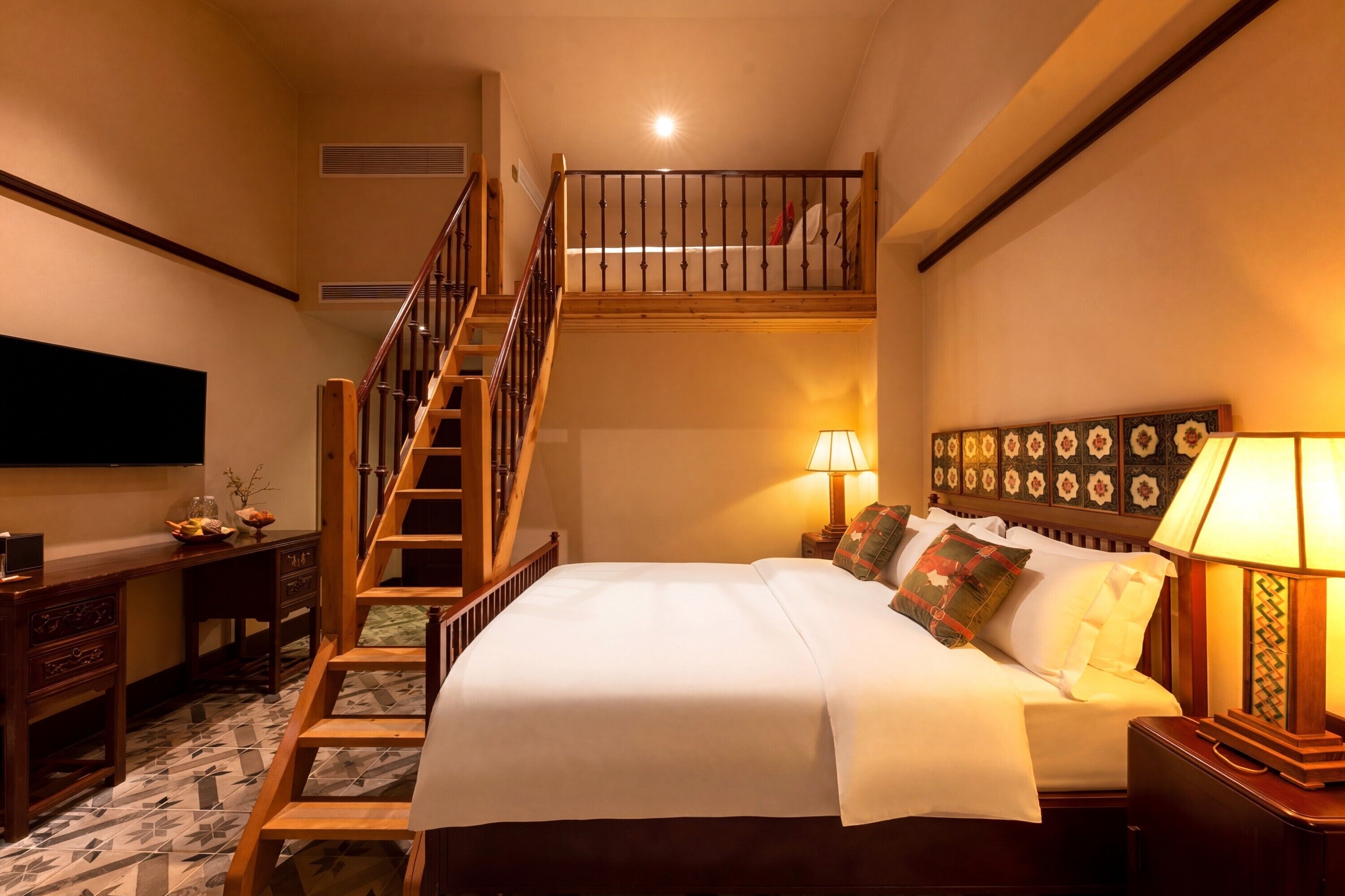 A hotel room with a bed, a nightstand, a lamp, a desk with a chair, a TV, and a staircase leading to an upper level.