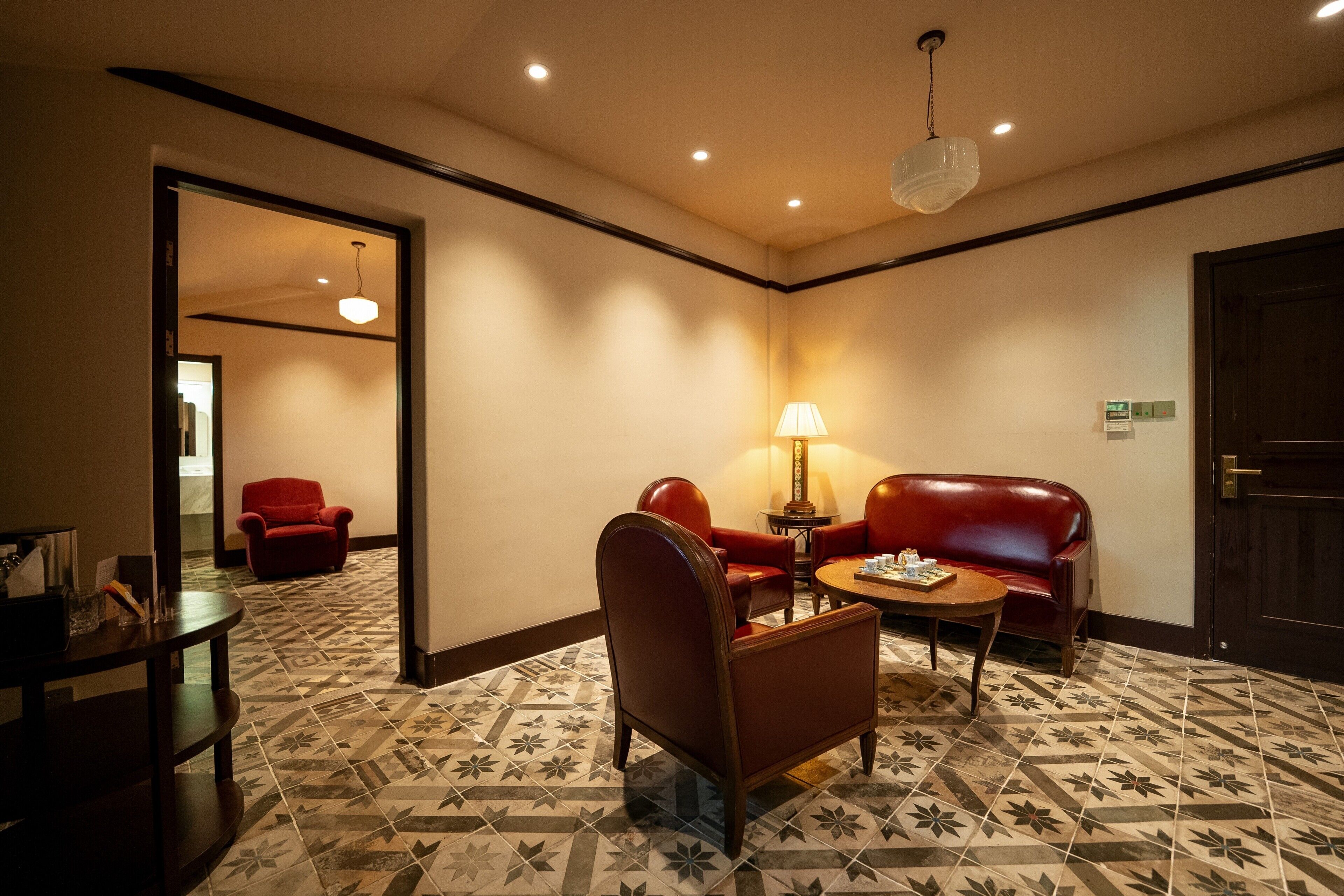 A hotel room with a patterned floor, a red leather sofa, a wooden coffee table, and a lamp.
