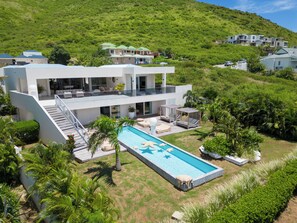 Ein modernes, zweistöckiges Haus mit Swimmingpool, umgeben von üppiger Vegetation und Hügeln.