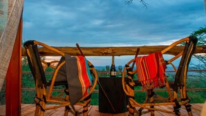 Two wooden chairs with woven seats and backs, a small table with a bottle and glasses, and a view of the landscape through a wooden structure. - Amani Eserian Ngorongoro (Ngorongoro)