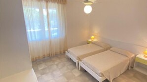 Een kamer met twee bedden, een plafondventilator, een raam met gordijnen en een bedlampje.