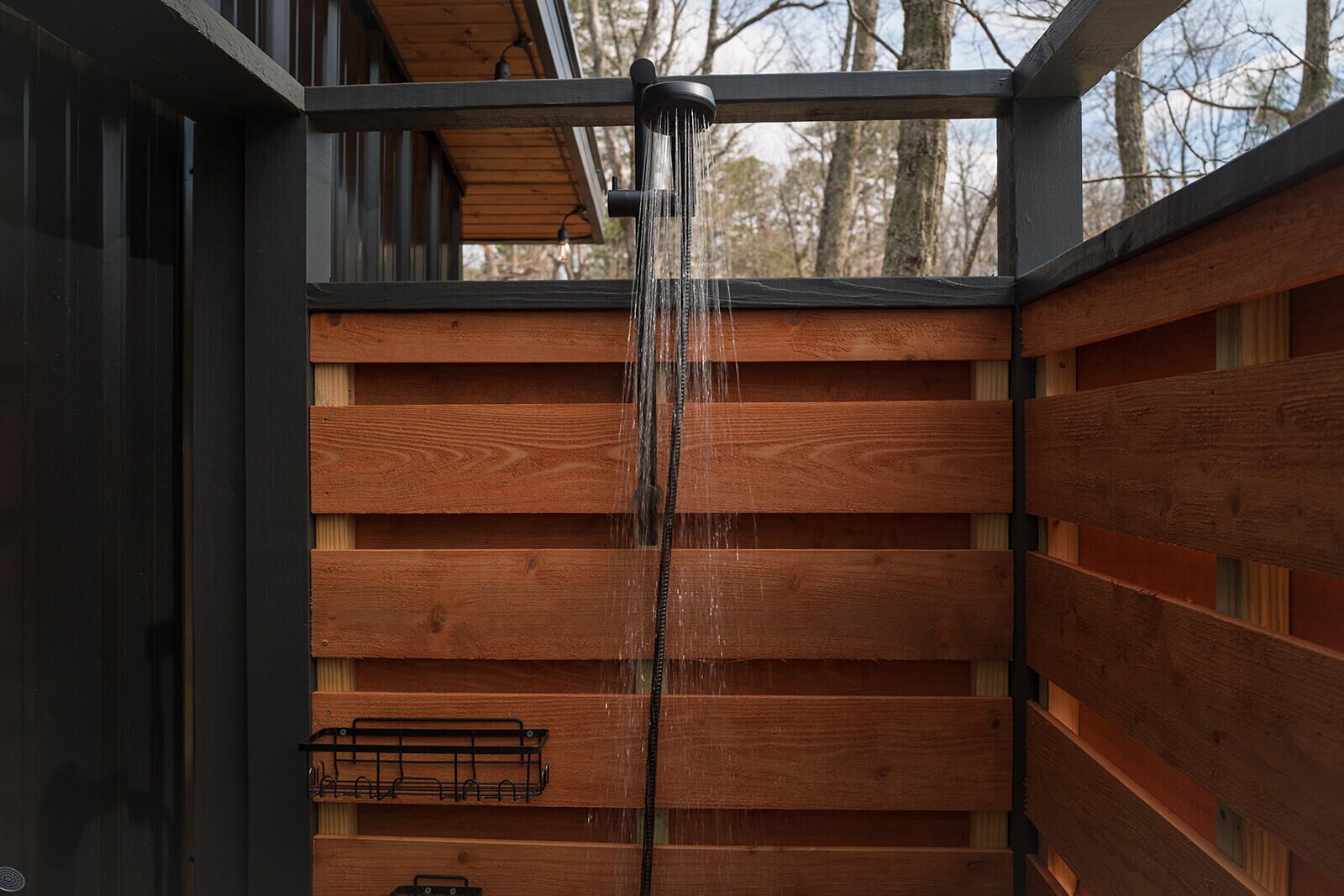 Una ducha exterior de madera con estructura metálica, un colgador metálico y vistas a árboles al fondo.