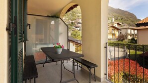 A balcony with a table and bench, a potted plant, and a view of a residential area with houses and a mountain. - Serenity Como Lake View Apartment With Pool (Tremezzina)