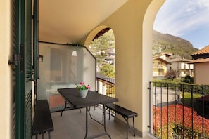A balcony with a table and bench, a potted plant, and a view of a residential area with houses and a mountain. - Serenity Como Lake View Apartment With Pool (Tremezzina)