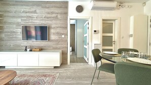 A modern living room with a flat-screen TV, a dining area with a round table and chairs, and a hallway leading to another room.
