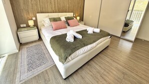 A modern bedroom with a large bed, a nightstand, a bedside lamp, a wooden wall, and a view through a sliding door.