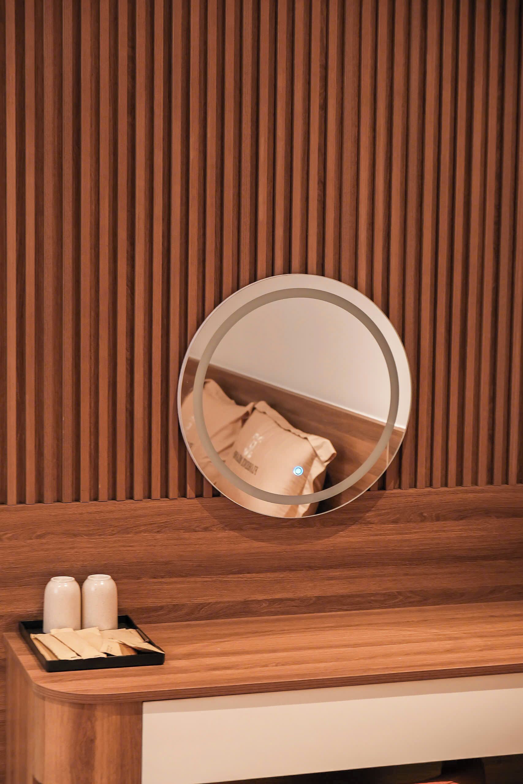 A wooden vanity with a round mirror, a tray with two vases, and a beige bag on the counter.