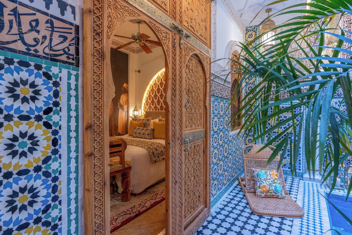 Una habitación ornamentada con detalles intrincados en azulejos, una cama con colcha estampada y una silla de mimbre con cojín.