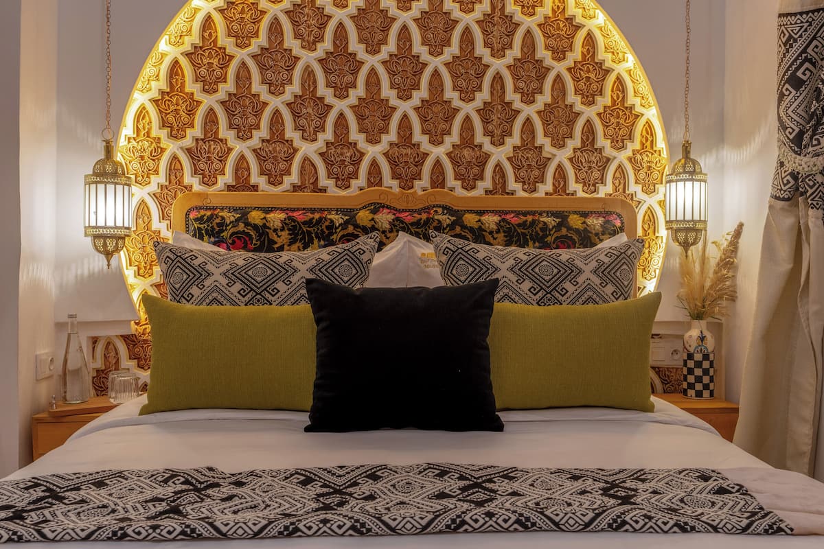 Una cama con ropa de cama estampada, un cabecero decorativo y dos lámparas fijadas a la pared.