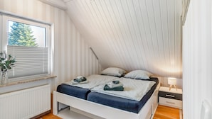 A bedroom with a sloped ceiling, a bed with pillows and folded towels, a bedside table with a lamp, and a window with blinds. - Holiday Home for 5 People (105 m²) in Südbrookmerland (Südbrookmerland)